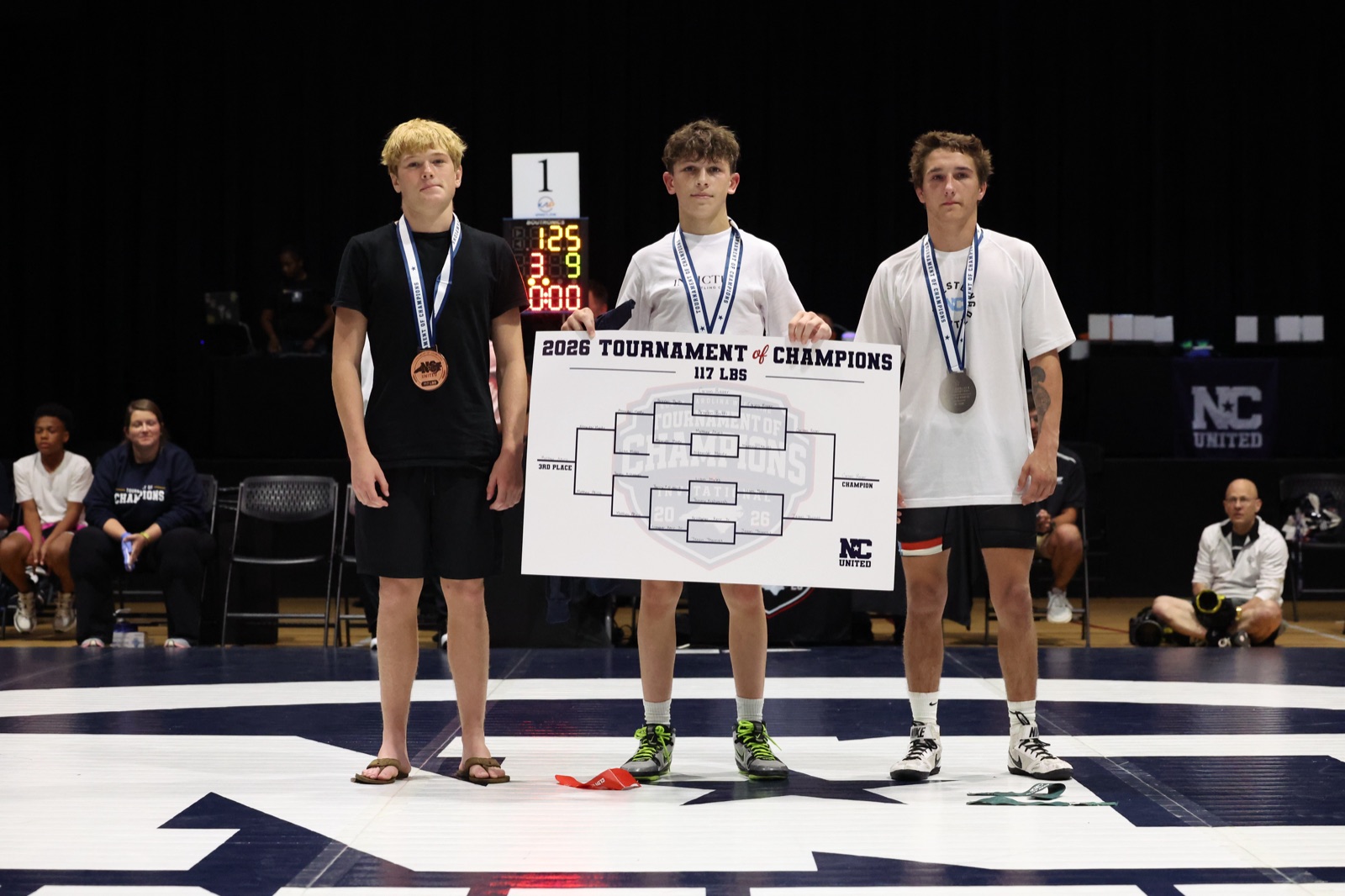 The 117 lb podium at the 2026 Tournament of Champions — the three placers on the mat, the champion holding the bracket